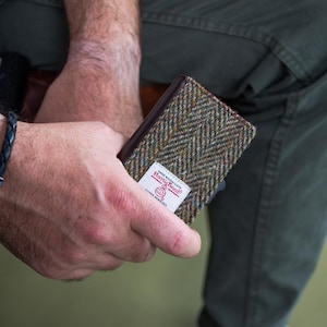 May include: A rectangular wallet with a brown leather exterior and a tweed fabric front. The tweed features a herringbone pattern in shades of green, blue, and brown. A white label with red text is visible on the front. The wallet is held in a person's hand.