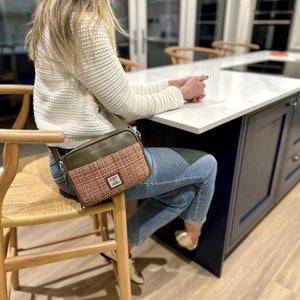 May include: A woman wearing a white sweater and blue jeans is sitting on a wooden stool. She is carrying a brown and pink tweed crossbody bag with the word "Maccessory" printed on the front.