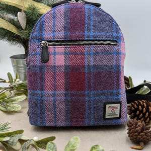 May include: A purple, pink, and blue plaid backpack with a black zipper and leather pull. The bag has a small black handle and a label on the front. The bag is sitting on a neutral surface with greenery and a small Christmas tree in the background.