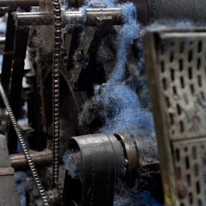 Op de afbeelding: Close-up van een vintage industri&euml;le machine met een kettingaandrijving en een riem. Blauwe vezels zijn om de tandwielen en rollen van de machine gewikkeld.