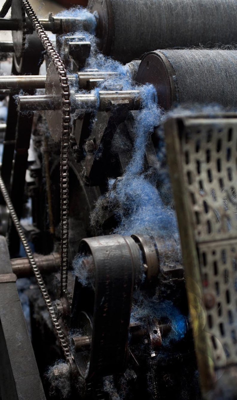 Op de afbeelding: Close-up van een vintage industri&euml;le machine met een kettingaandrijving en een riem. Blauwe vezels zijn om de tandwielen en rollen van de machine gewikkeld.