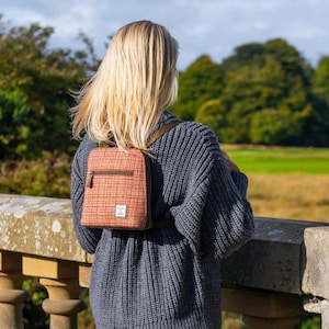 Puede incluir: Una pequeña mochila de tweed naranja y marrón con cremallera y correas marrones. La mochila se lleva sobre un suéter gris. El fondo presenta un muro de piedra, hierba verde y árboles.