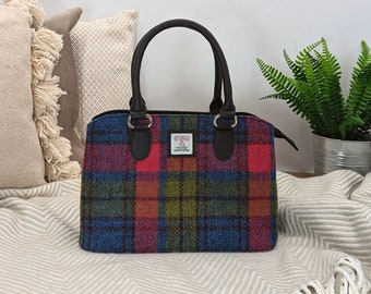 May include: A colorful plaid handbag with a structured shape. The bag features a mix of red, blue, green, and brown tones. It has dark brown handles and accents, and a small label is visible on the front. The bag is sitting on a cream-colored blanket.
