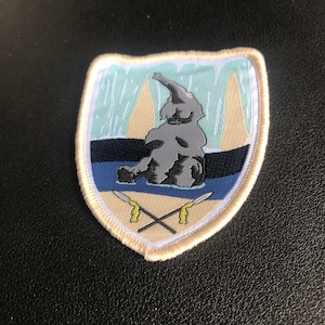 Peut inclure: Un écusson brodé beige avec une bordure blanche. L'écusson représente un éléphant gris en dessin animé avec une longue trompe debout dans une étendue d'eau bleue. L'éléphant est devant une cascade beige et bleue. Deux lances croisées avec des pointes jaunes se trouvent au bas de l'écusson.