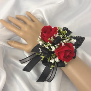 May include: A wrist corsage featuring two red roses, accented with small white flowers and green sprigs. Black ribbon and a black glittery base complete the design. The corsage is designed to be worn on the wrist.