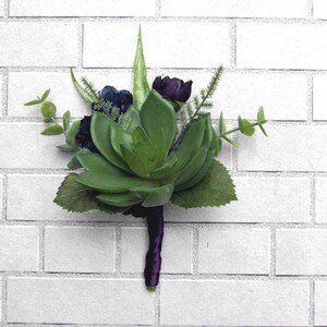May include: A succulent boutonniere with purple flowers and green leaves, attached to a purple ribbon.