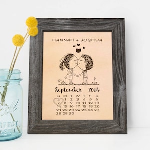 May include: A framed print with a hand-drawn illustration of a couple kissing on a calendar page for September 2014. The names Hannah and Joshua are printed at the top of the print.