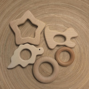 May include: Five wooden teethers in different shapes, including a star, a dinosaur, a ring, and two other shapes. The teethers are all natural wood color and are arranged on a wooden surface.