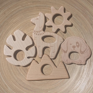 May include: A collection of wooden baby teethers in various shapes, including a llama, a sun, a leaf, a dog, and a mountain. Each teether is light brown and features a smooth, unfinished surface with a hole for easy gripping.