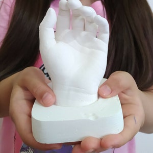 May include: A white plaster hand cast, a keepsake of a child's hand, is held by a person. The cast is on a white square base.