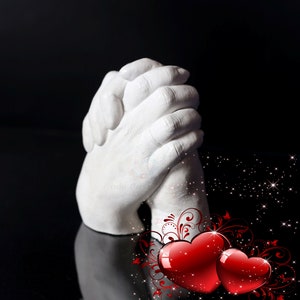 May include: White plaster hand cast of two hands clasped together, with a red heart and a red and white floral design in the background.