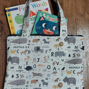 May include: A white tote bag with a zipper closure and black handles. The bag is printed with a colorful alphabet and animal illustrations. The bag is filled with three children's books: "First Words", "1, 2, 3 Eat With Me", and "Alphaprints".