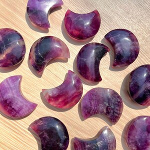 May include: A collection of 12 purple and pink crescent moon shaped fluorite crystals. The crystals are polished and have a smooth, shiny surface.