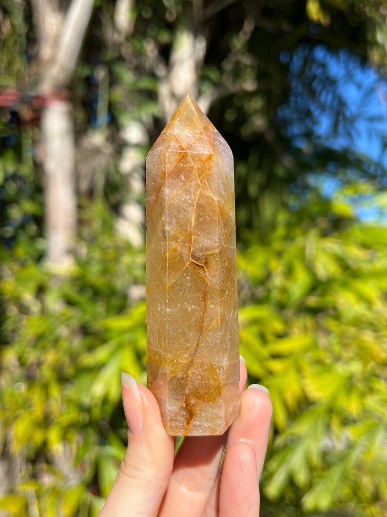 May include: A clear quartz crystal point with golden inclusions. The crystal is faceted and has a pointed tip. It is held in a hand against a blurred green background.