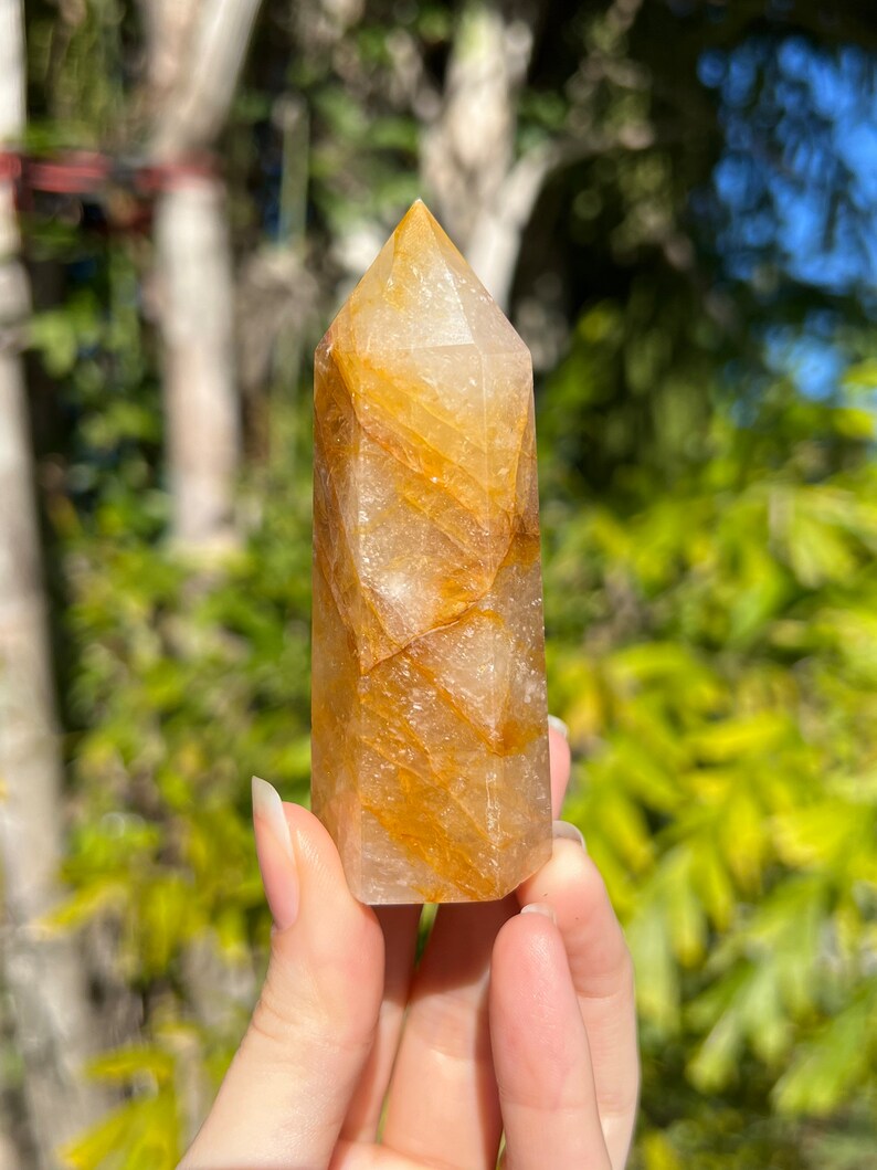 May include: A clear quartz crystal point with golden inclusions. The crystal is faceted and has a pointed tip. It is approximately 5 cm tall.