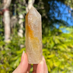 May include: A clear quartz crystal point with a golden yellow hue. The crystal is faceted and has a pointed tip. It is held in a hand against a blurred background of green foliage.