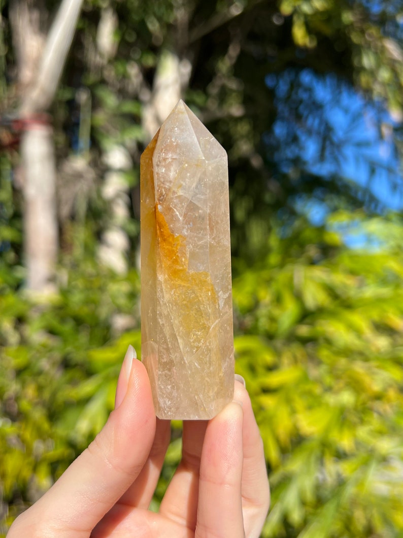 May include: A clear quartz crystal point with a golden yellow hue. The crystal is faceted and has a pointed tip. It is held in a hand against a blurred background of green foliage.