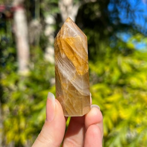 May include: A golden citrine crystal point with white streaks and a pointed tip. The crystal is held in a hand.
