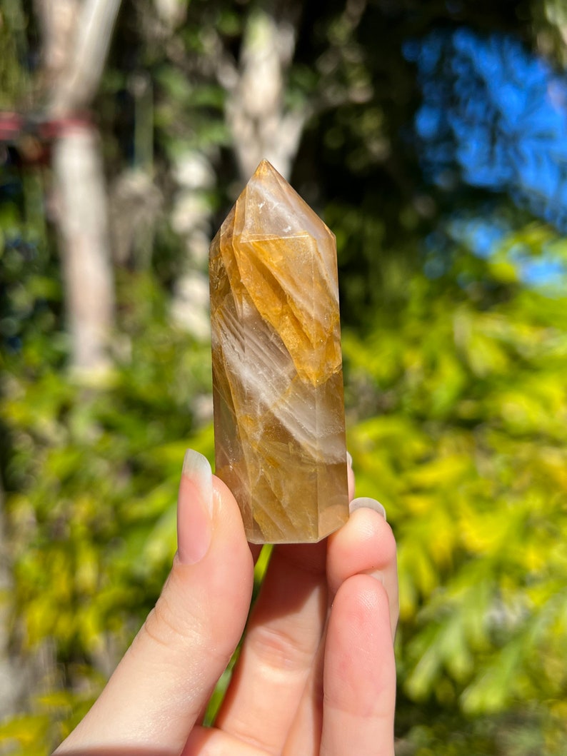 May include: A golden citrine crystal point with white streaks and a pointed tip. The crystal is held in a hand.