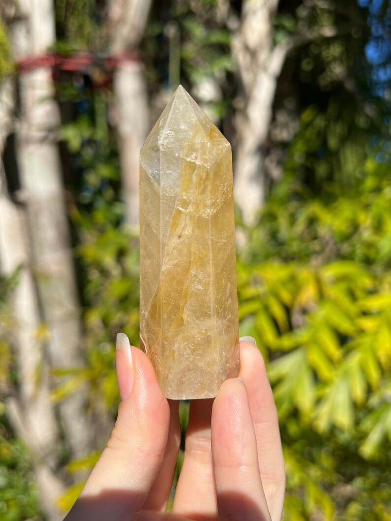 May include: A clear quartz crystal point with golden inclusions. The crystal is faceted and has a pointed tip. It is a natural crystal and has a unique shape and colour.
