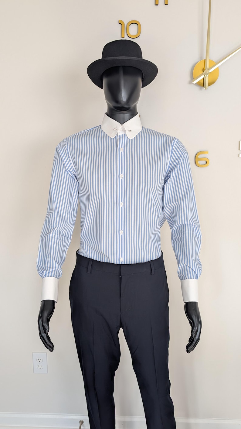 May include: A mannequin wearing a black hat, a blue and white striped shirt with a white collar and cuffs, and black pants. The shirt has vertical stripes. The mannequin is wearing black gloves.