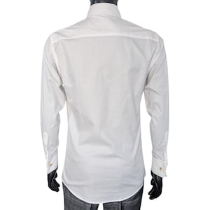 Op de afbeelding: Een wit overhemd met lange mouwen en gouden manchetknopen wordt op een mannequin getoond. Het overhemd wordt gecombineerd met een grijze geruite broek. De armen en handen van de mannequin zijn zwart.