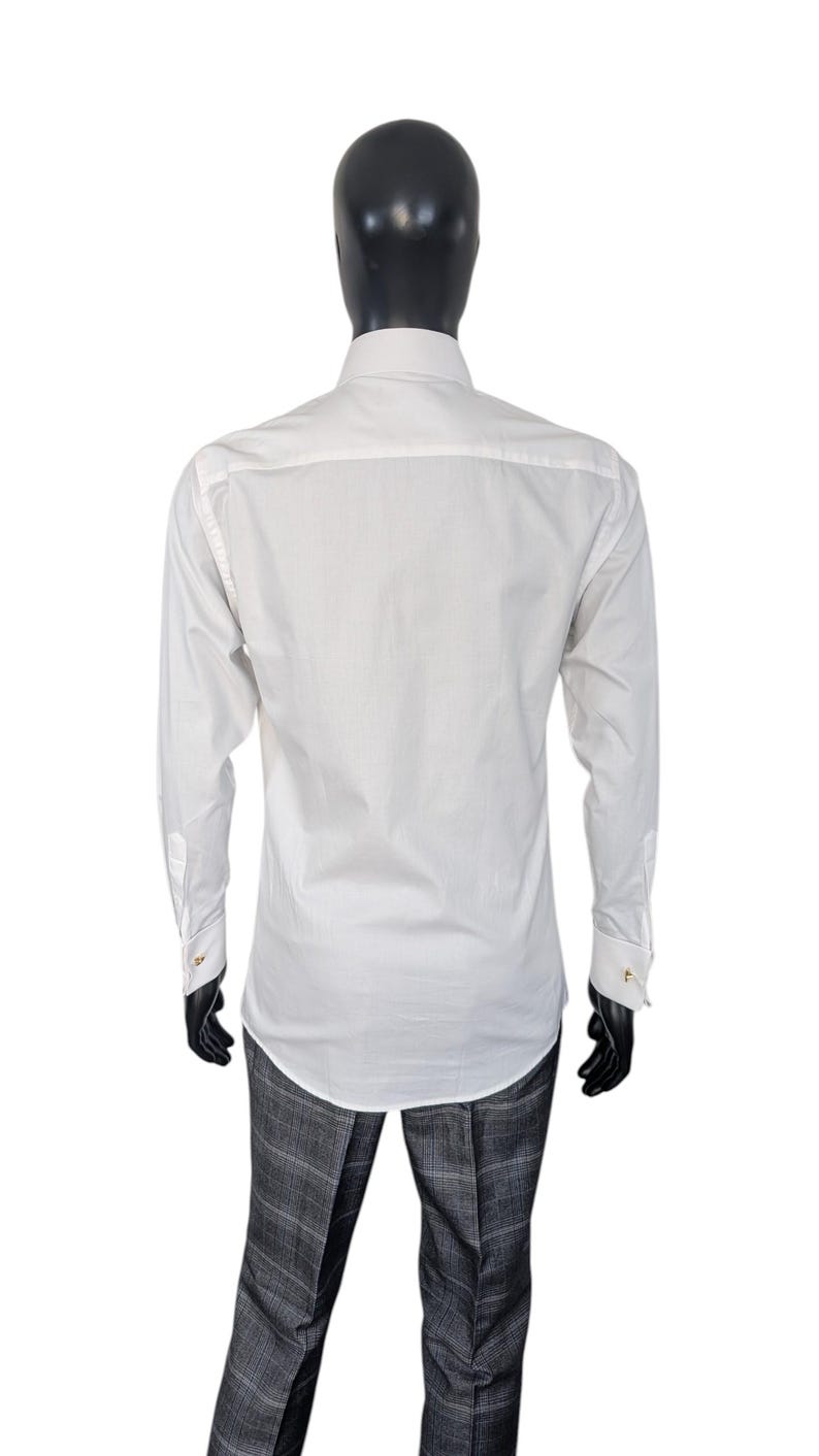 Op de afbeelding: Een wit overhemd met lange mouwen en gouden manchetknopen wordt op een mannequin getoond. Het overhemd wordt gecombineerd met een grijze geruite broek. De armen en handen van de mannequin zijn zwart.