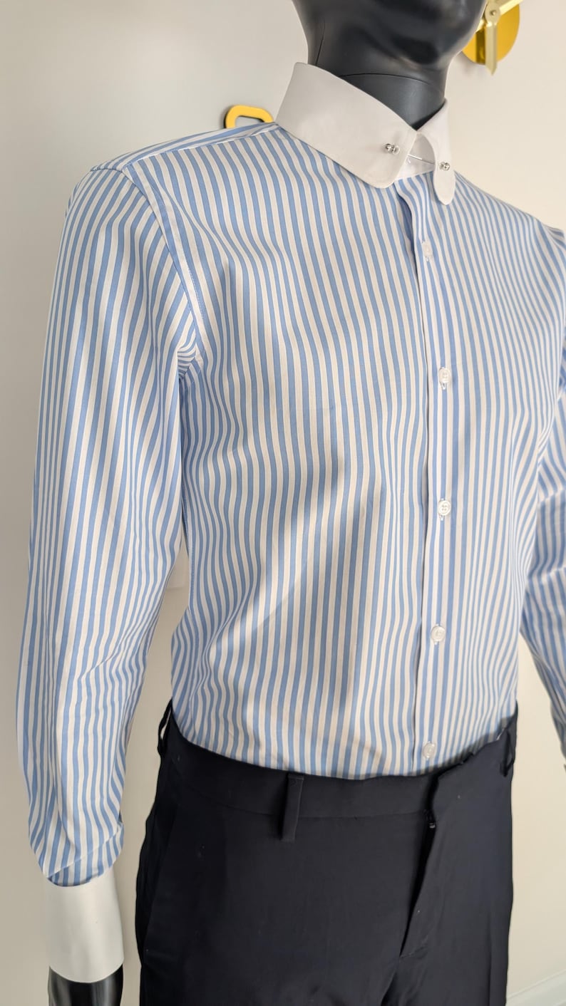 May include: A blue and white striped dress shirt with a white collar and cuffs. The shirt is buttoned up and paired with black dress pants. The shirt is on a mannequin.