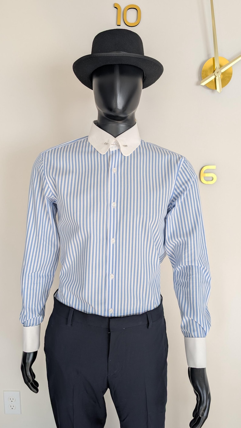 May include: A black hat sits atop a mannequin wearing a blue and white striped shirt with a white collar and cuffs. The mannequin is also wearing dark blue pants. A gold clock is visible in the background.