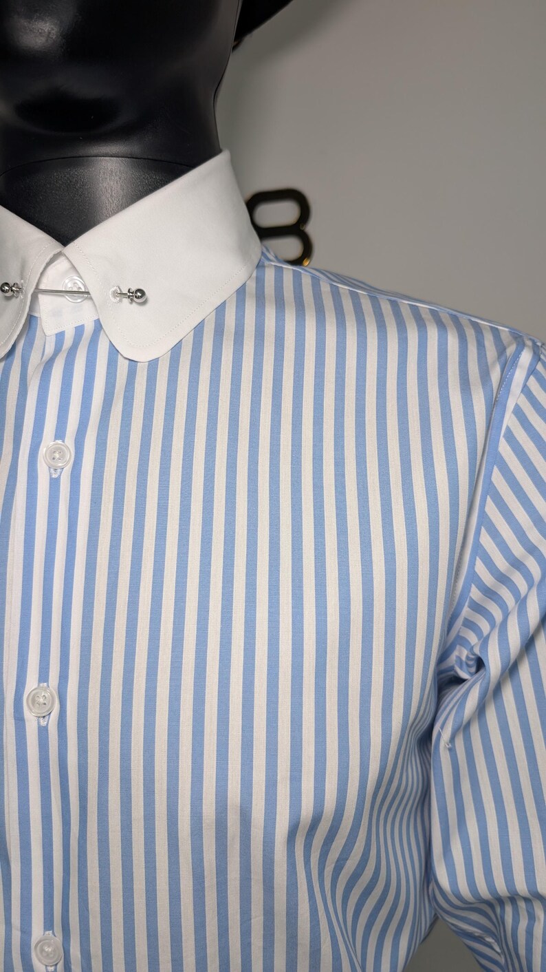 May include: A blue and white striped dress shirt with a white collar and buttoned cuffs. The shirt has a classic design with vertical stripes and a collar pin.