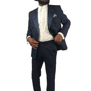 Op de afbeelding: Een marineblauw dubbelbreasted pak met een satijnen revers, gecombineerd met een wit gerimpeld overhemd, een zwarte fedora en zwarte schoenen. Een pochet voegt een vleugje detail toe aan dit formele ensemble.
