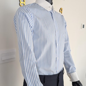 May include: A mannequin wearing a blue and white striped dress shirt with a white collar and cuffs. The shirt is buttoned up and paired with black pants. A black hat sits atop the mannequin's head. Gold cufflinks are visible.