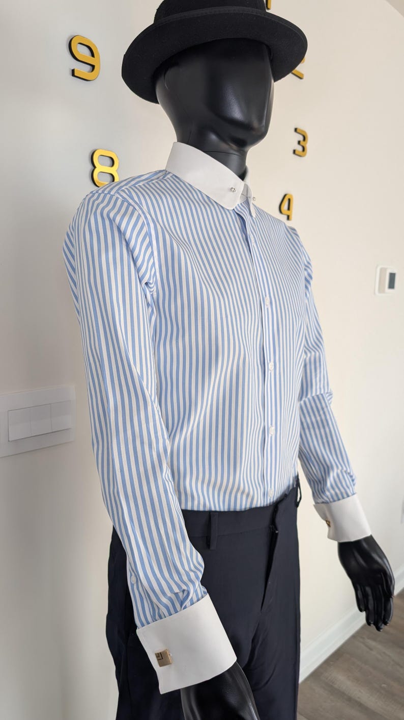 May include: A mannequin wearing a blue and white striped dress shirt with a white collar and cuffs. The shirt is buttoned up and paired with black pants. A black hat sits atop the mannequin's head. Gold cufflinks are visible.