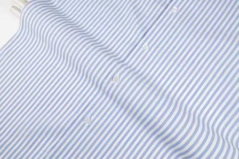 May include: Close-up of a light blue and white striped fabric. The fabric is a woven material with vertical stripes. The image shows the fabric's texture and the subtle variations in the stripes. Several white buttons are visible.