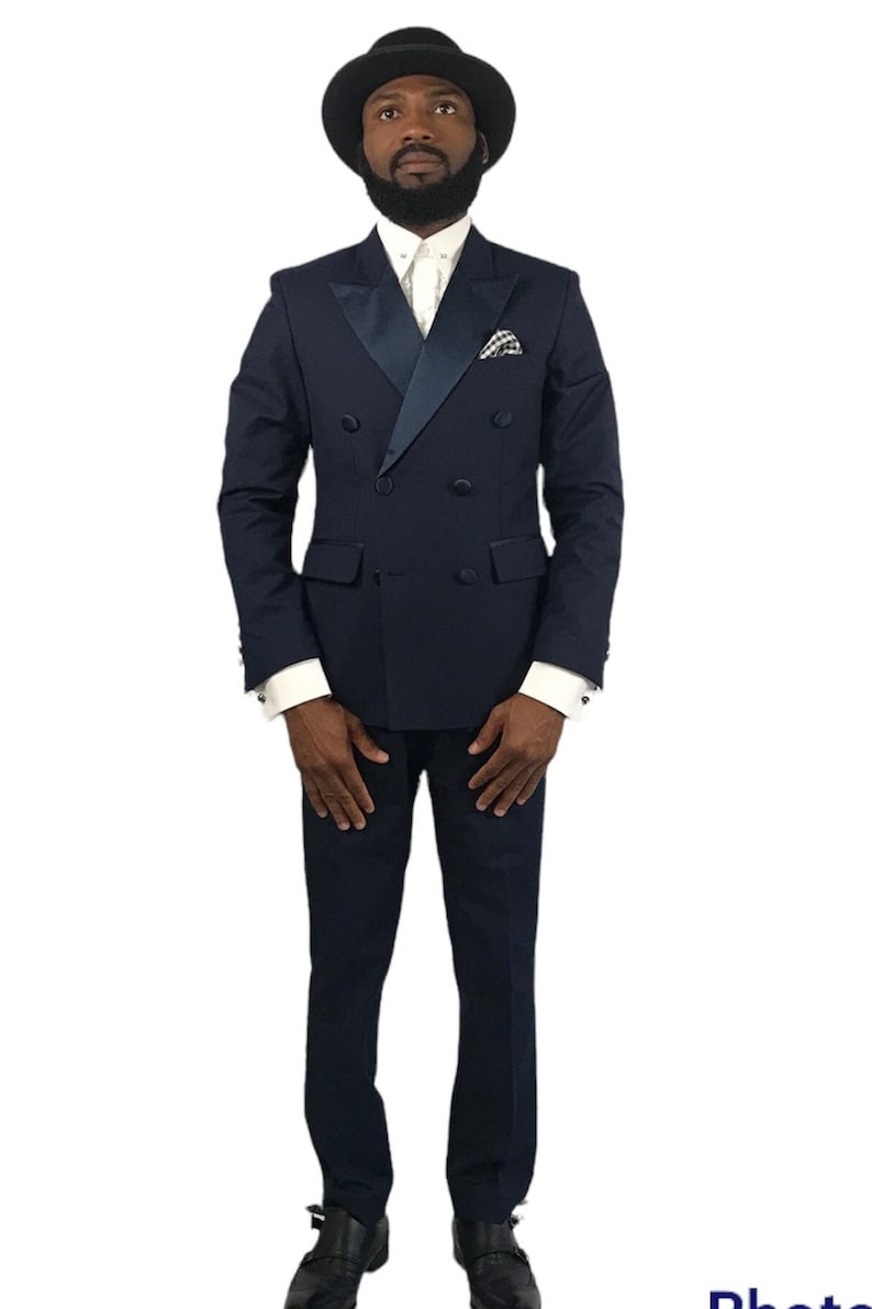 Op de afbeelding: Een marineblauw dubbelbreasted pak met een satijnen revers, gecombineerd met een wit overhemd en een zwarte hoed. Het pak heeft een pochet en wordt gedragen met zwarte schoenen. Een formele outfit.