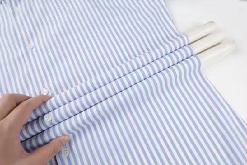 May include: A blue and white striped button-down shirt. The shirt is made of a lightweight fabric and features vertical stripes. The buttons are white. The shirt is folded over a white hanger.