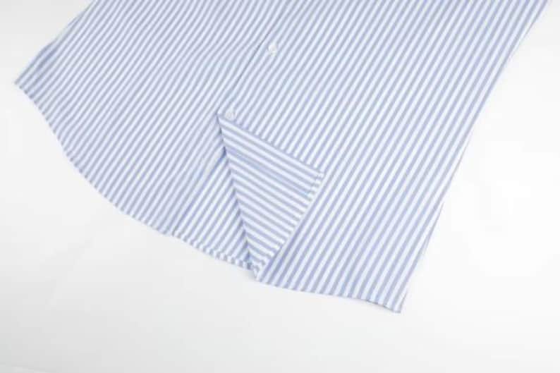 May include: Close-up of a light blue and white striped shirt. The fabric is folded, revealing a button and the edge of the shirt. The pattern consists of vertical stripes, creating a classic and clean look. The shirt is made of a lightweight material.