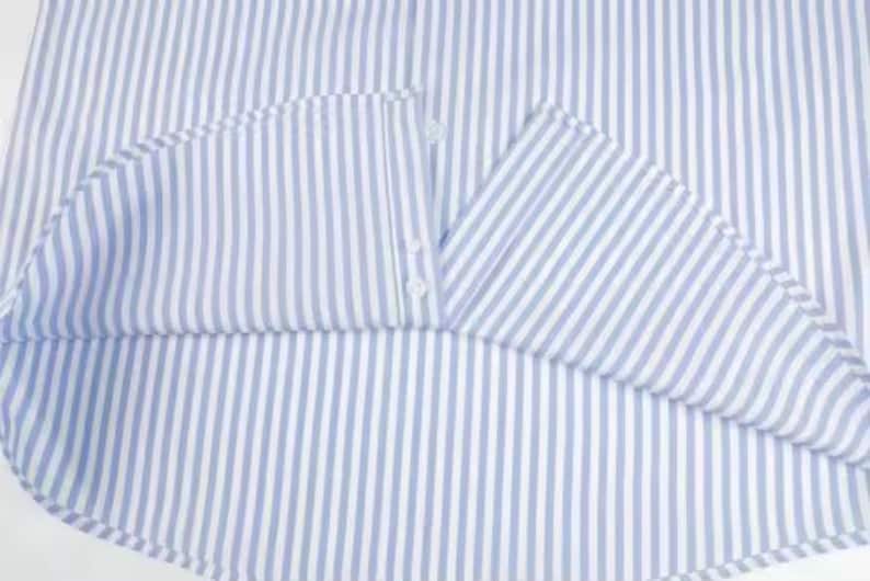 May include: Close-up of a light blue and white striped shirt. The fabric is folded, revealing the inside of the collar and the button closure. The shirt's pattern is a classic vertical stripe.