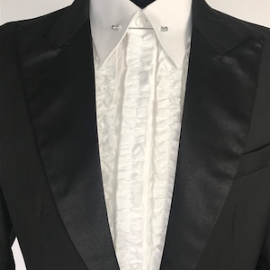 Op de afbeelding: Een zwarte smokingjas met satijnen revers, gecombineerd met een wit gerimpeld overhemd. Het overhemd heeft een klassieke kraag en wordt vastgezet met een kraagspeld. De formele kleding wordt op een mannequin gepresenteerd.