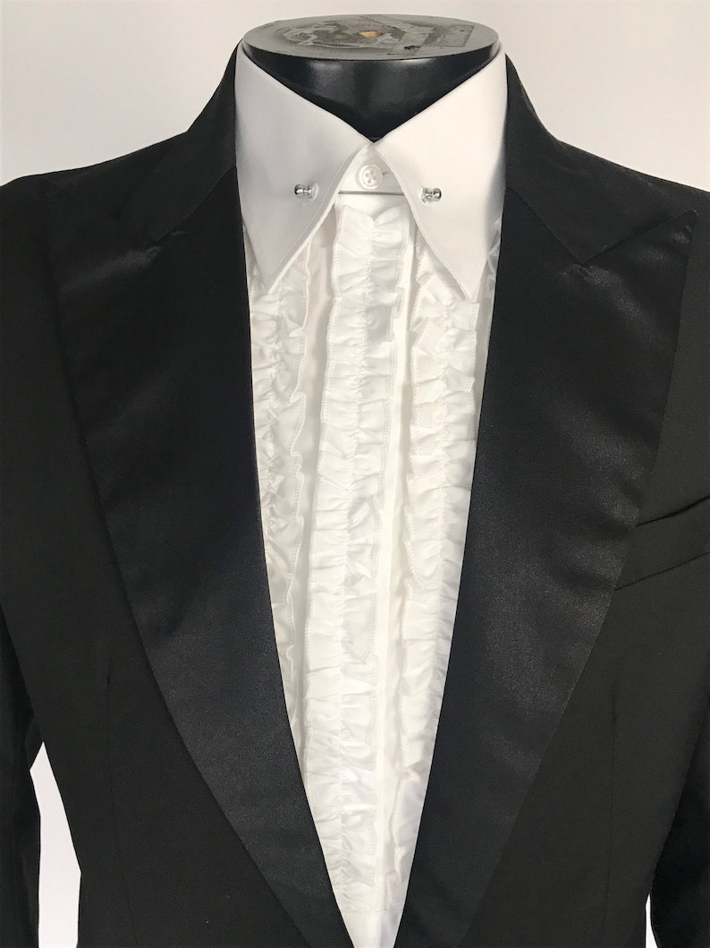 Op de afbeelding: Een zwarte smokingjas met satijnen revers, gecombineerd met een wit gerimpeld overhemd. Het overhemd heeft een klassieke kraag en wordt vastgezet met een kraagspeld. De formele kleding wordt op een mannequin gepresenteerd.