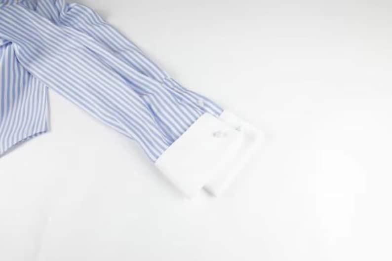 May include: Close-up of a blue and white striped dress shirt sleeve with a white cuff. The cuff has two buttons. The shirt is made of a lightweight fabric and is partially visible on a white background.