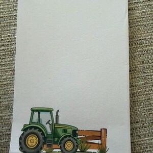 May include: A white notepad with a green tractor illustration at the bottom. The tractor has yellow wheels and is next to a brown fence and green grass. The notepad is on a textured surface.