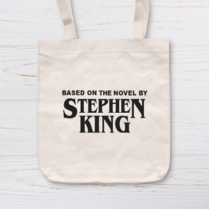 Peut inclure: Un sac fourre-tout crème avec le texte noir "BASED ON THE NOVEL BY STEPHEN KING". Le sac a deux anses crème et est fait d'un tissu robuste. Le design est simple, avec des lettres noires et audacieuses, faciles à lire.