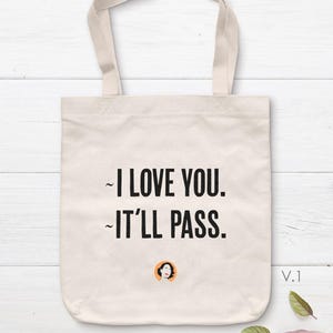 Pode incluir: Uma sacola de lona bege com duas alças, apresentando o texto impresso em preto "I LOVE YOU. IT'LL PASS." e um pequeno ícone circular laranja contendo uma ilustração estilizada de uma pessoa com cabelo escuro e lábios vermelhos.