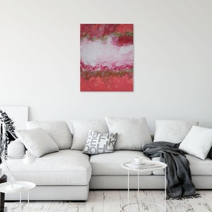 May include: Abstract art piece with a red, white, and gold color scheme. The artwork is a vertical rectangle, with a layered effect of colors. The piece is displayed on a white wall above a white couch.
