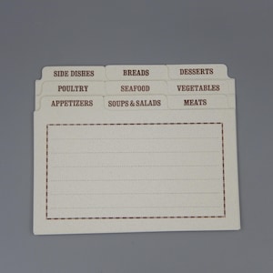 May include: A white recipe card holder with brown tabs labeled with different food categories: Side Dishes, Poultry, Appetizers, Breads, Seafood, Soups & Salads, Desserts, Vegetables, and Meats.