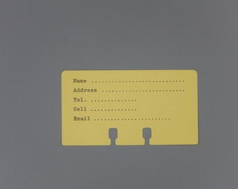 May include: A yellow index card with the words "Name", "Address", "Tel", "Cell", and "Email" printed on it. The card has two holes punched in the top for hanging.