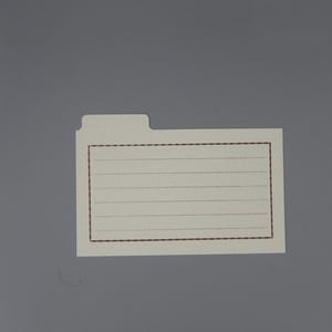 May include: A cream-colored file folder label with a brown dotted border and lines for writing. The label has a tab at the top and is set against a gray background.