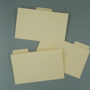 May include: A collection of cream-colored file folders with attached tabs, arranged on a gray surface. The folders are rectangular, with a stack of papers visible in one. The tabs are positioned at the top.