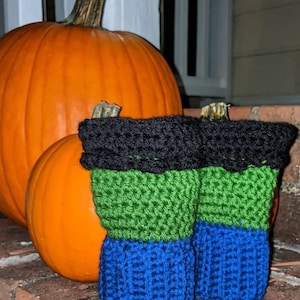 May include: A pair of crocheted leg warmers in green, blue, and black. The leg warmers are sitting in front of two pumpkins.